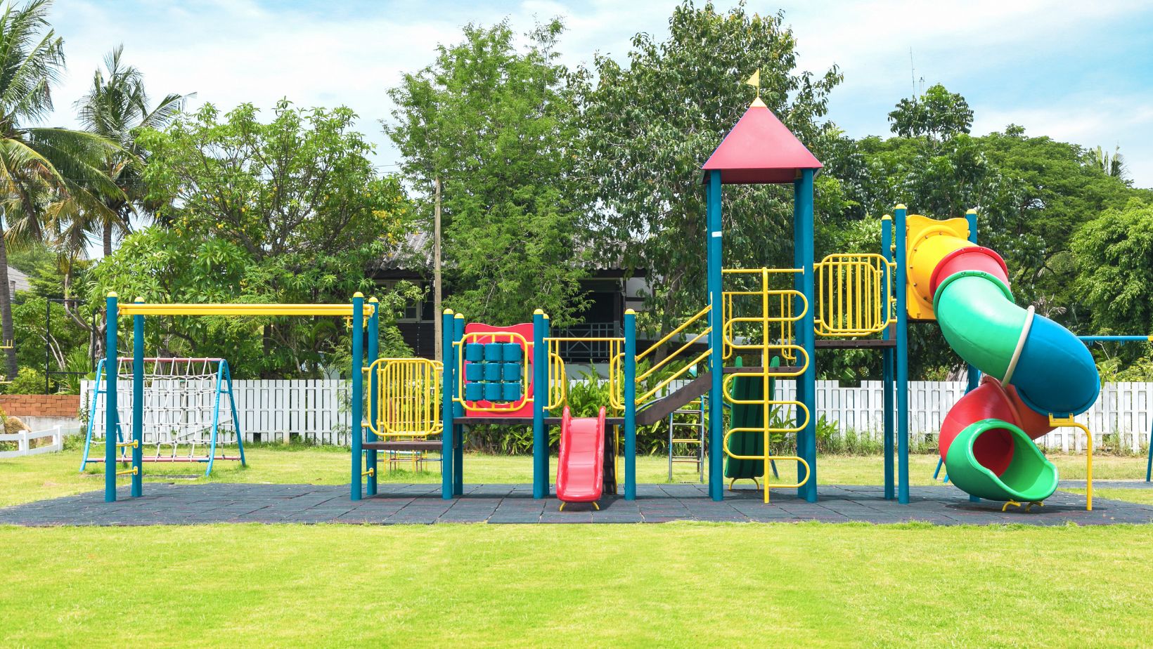 A colorful playground with a slide, swings, and climbing structures on a grassy field. Trees and a white fence are in the background under a blue sky.