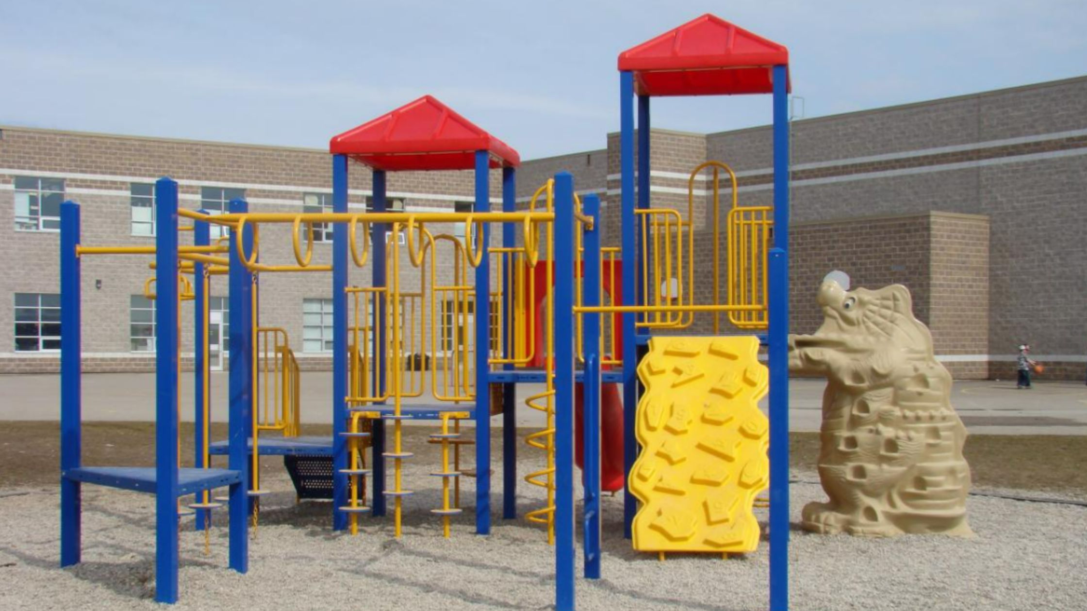 Playground with colorful climbing structures, slides, and ladders in yellow and blue. Background features a brick school building. Sunny day.